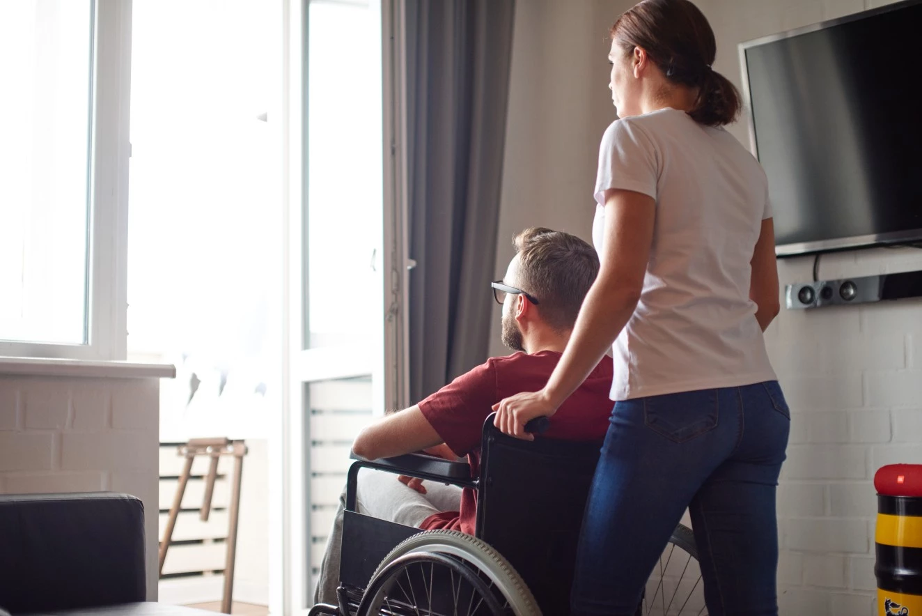 Disabled man in wheelchair at home with support worker behind him both staring out window